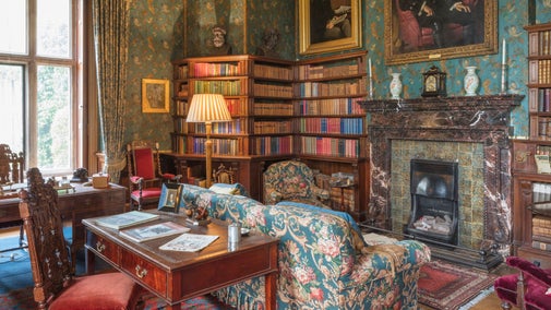 A library with ornate blue wallpaper, a marble fireplace in the centre, and dark wood bookshelves. There are a blue sofa and armchair and wooden desk and chair in the room.